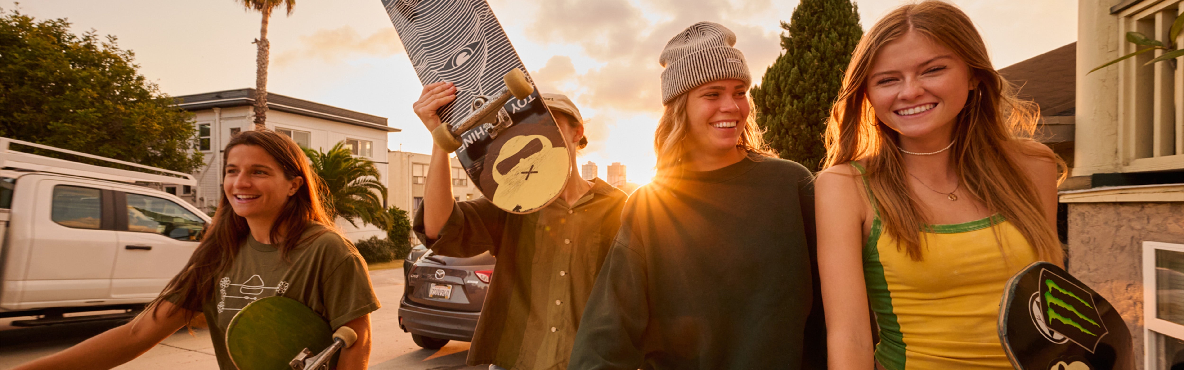 Four friends smiling and skating at sunset, holding boards from the SB Sun Care Collection – Sun Bum.