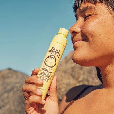 Smiling person holding Sun Bum Mist 45 sunscreen spray, ideal for quick, refreshing sun protection - Original SPF Collection - Sun Bum