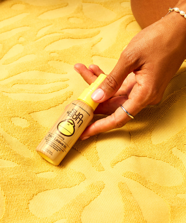 A hand holding Sun Bum's 3 In 1 Leave In hair treatment bottle against a vibrant yellow textured beach towel background, showcasing the multifunctional hair care product from Sun Bum