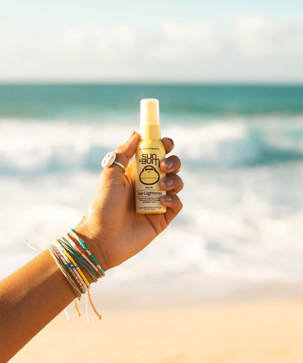 Hand holding Sun Bum Blonde Hair Lightener spray bottle against a beachy ocean backdrop, adorned with colorful friendship bracelets, showcasing the natural hair lightening product by Sun Bum
