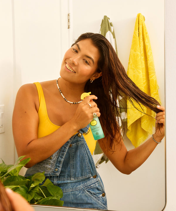 Person in yellow tank top and denim overalls spraying Heat Protector hair product in bathroom, demonstrating its use with beach-inspired styling spray that protects against heat damage and reduces frizz - by Sun Bum