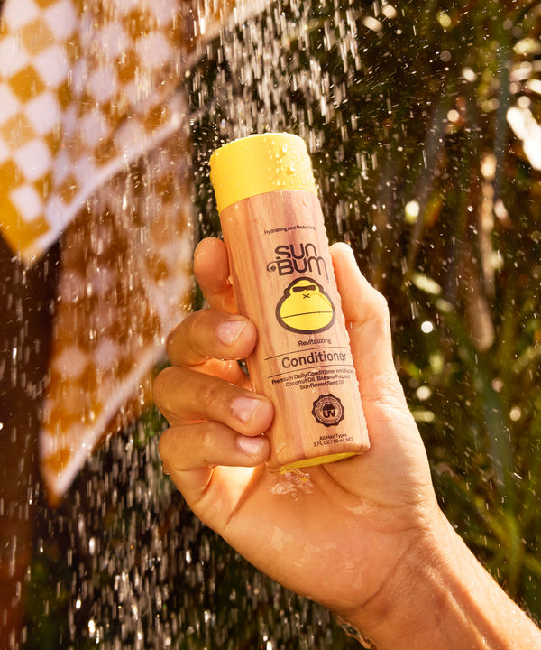 A hand holding Sun Bum Revitalizing Conditioner bottle with yellow cap against sparkling water droplets and golden sunlight, showcasing the hydrating haircare product by Sun Bum
