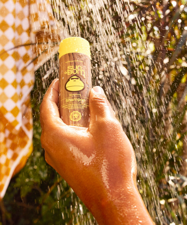 Revitalizing Shampoo bottle held up against sparkling water droplets in sunlight, showcasing the wooden-toned packaging with yellow cap and monkey logo. Natural outdoor setting with bokeh effects and warm golden tones. By Sun Bum.