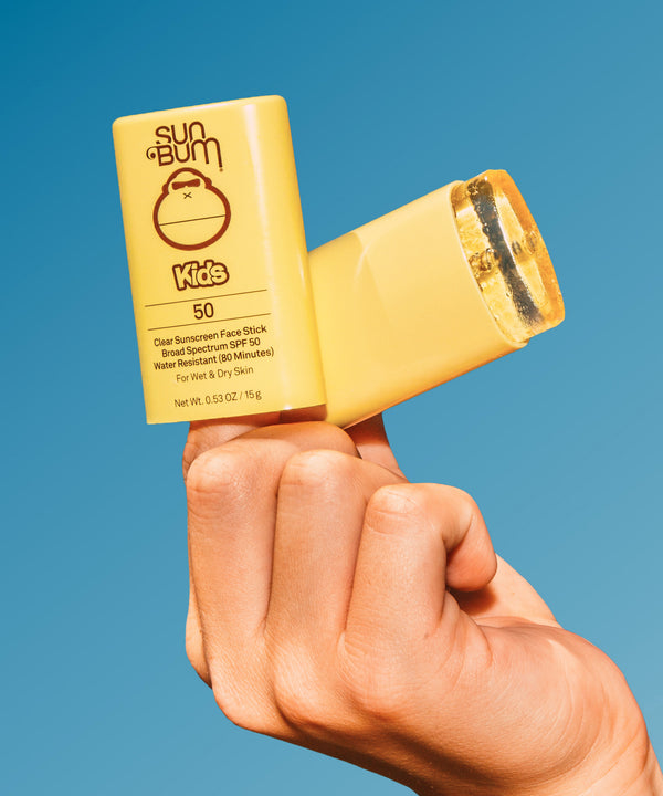 Hand holding a Sun Bum Kids SPF 50 Clear Sunscreen Face Stick against a blue sky. Sun Bum,
