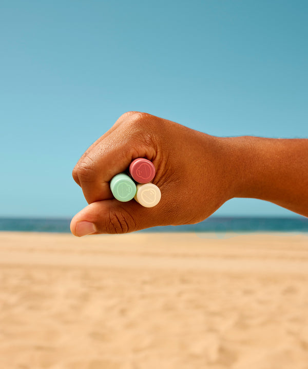 Hand holding three colorful CocoBalm Lip Balm tubes against a beachy background with turquoise sky and sandy shore, showcasing the 3-pack bundle by Sun Bum