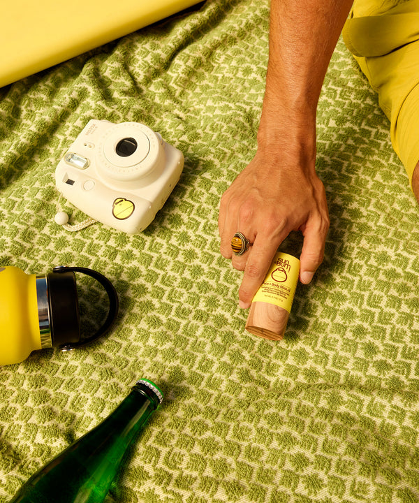 Hand holding Original Face and Body Stick 40 on green blanket with camera, bottle, and yellow accessories, perfect for on-the-go SPF protection. Sun Bum