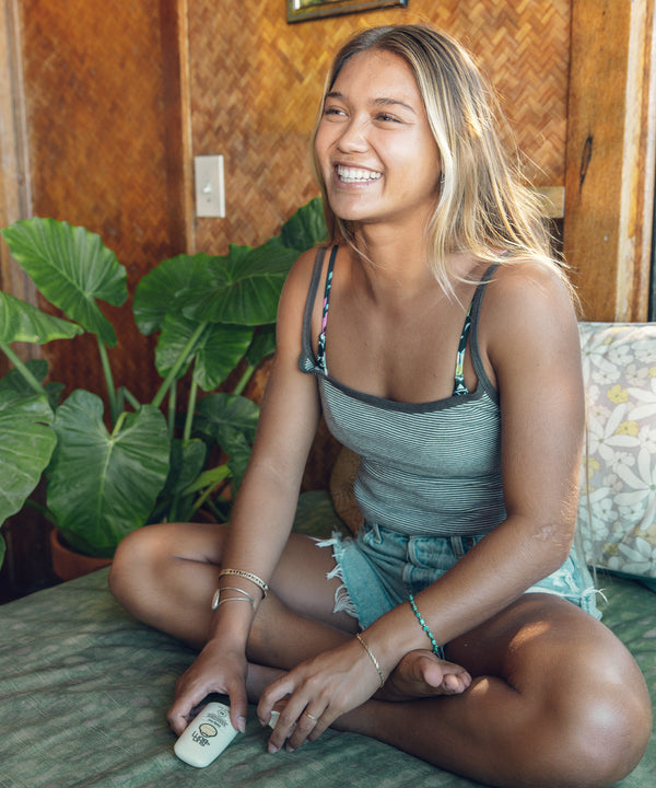 Woman smiling while holding a tube of Daily 50 Face Gel, sitting comfortably in a room with tropical plants. Sun Bum.