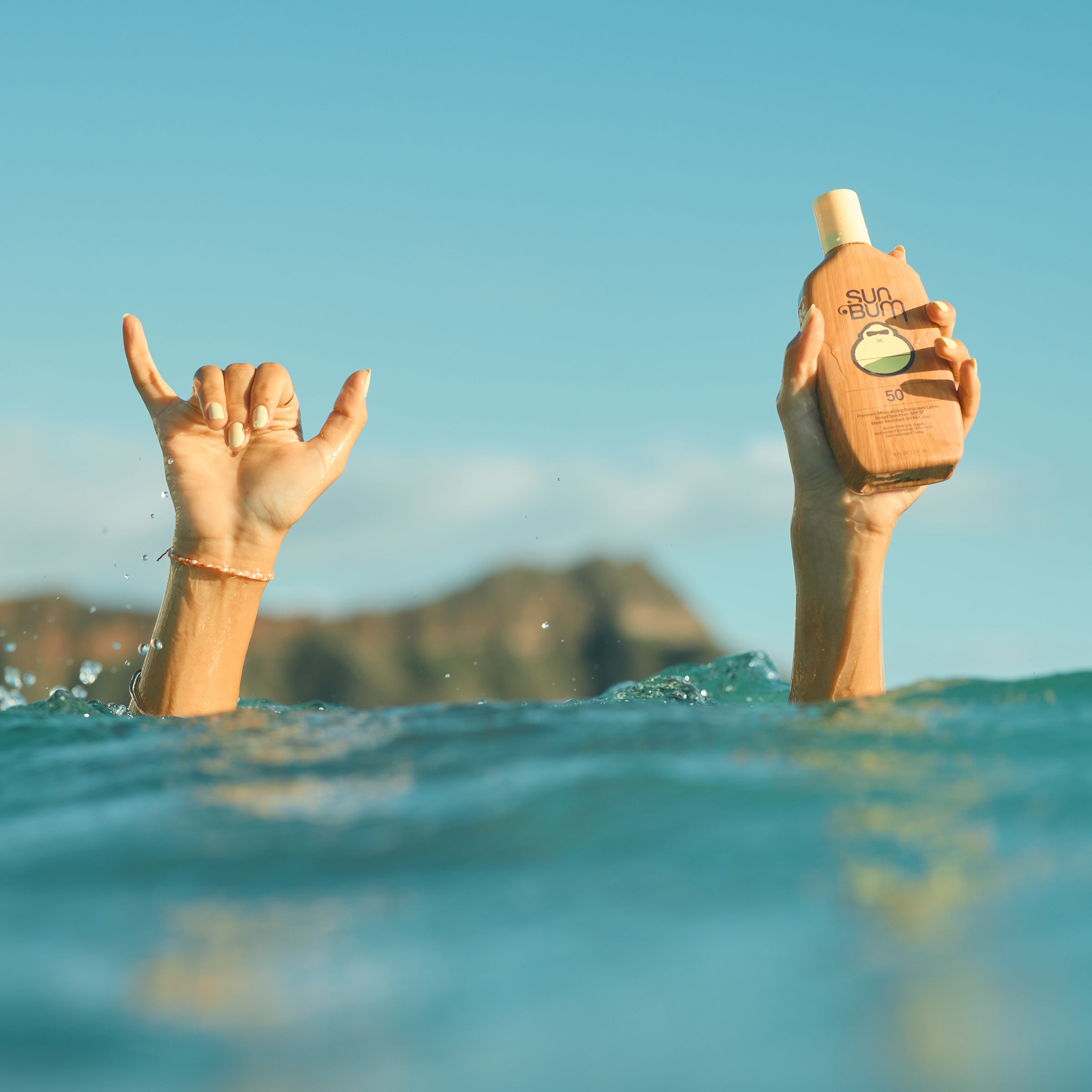 Hands rise from ocean with a shaka sign and Sun Bum SPF 50 lotion, highlighting water-resistant sunscreen - Original SPF Collection - Sun Bum