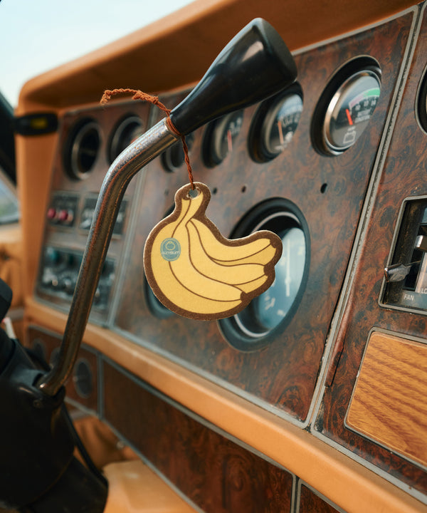 Yellow banana-shaped car air freshener hanging from vintage vehicle dashboard with wooden trim and classic gauges. Banana Air Freshener by Sun Bum brings summer vibes to your daily drive.