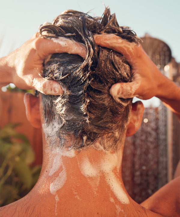 Person massaging Revitalizing Detox Scalp Scrub into wet hair, showing foamy lather and exfoliating action against a sunny outdoor backdrop. Natural sunlight highlights the rich texture of the product working into the scalp - Sun Bum