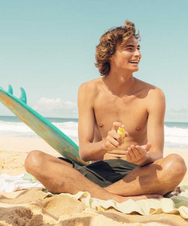 Shirtless man sitting on the beach, smiling and applying SPF 45 Sunscreen Face Mist with a surfboard in the background. Sun Bum.