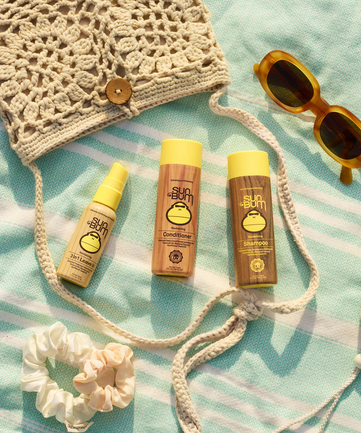 Three Sun Bum hair care products including the 3 In 1 Leave In spray, conditioner, and shampoo displayed on a mint striped beach towel with vintage sunglasses, crochet bag, and cream scrunchie for a summer-ready hair care routine - by Sun Bum