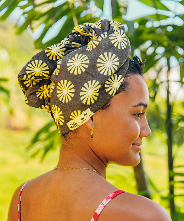 Side profile showing the Air Dry Cream & Hair Towel Kit in action - featuring a charcoal gray towel with yellow sunburst pattern wrapped elegantly around wet hair, photographed outdoors against lush tropical foliage in natural sunlight - by Sun Bum