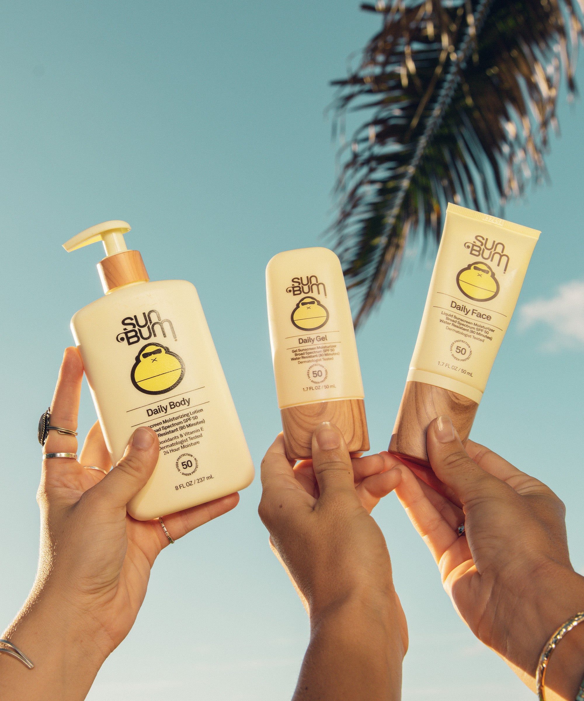 Three hands holding bottles of Sun Bum Daily 50 Body Lotion, enriched with Vitamin E and Antioxidants, outdoors against a sunny sky with a palm tree.