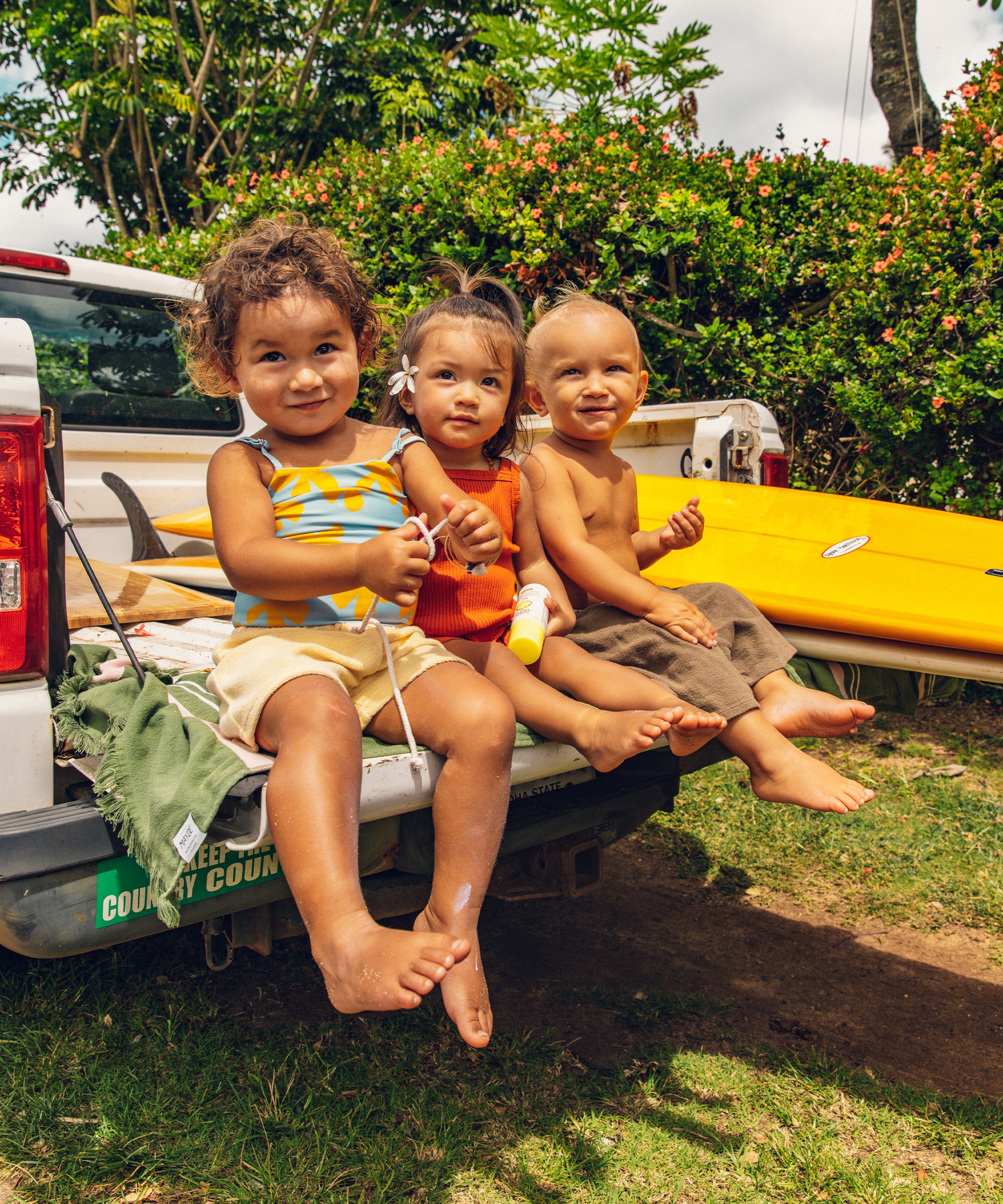 Three happy toddlers sitting on a truck tailgate with surfboards, holding Baby Bum Mineral SPF 50 Roll-On Sunscreen by Baby Bum