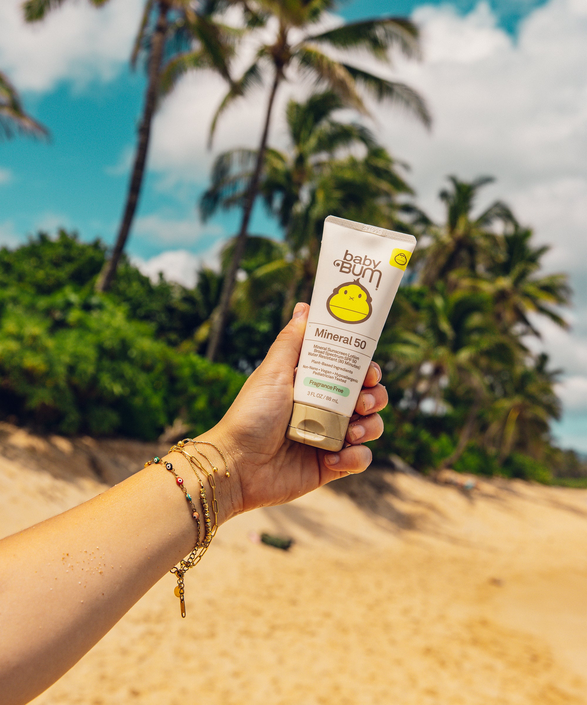Hand holding Baby Bum Mineral SPF 50 Sunscreen Lotion with beach background for sun protection by Baby Bum