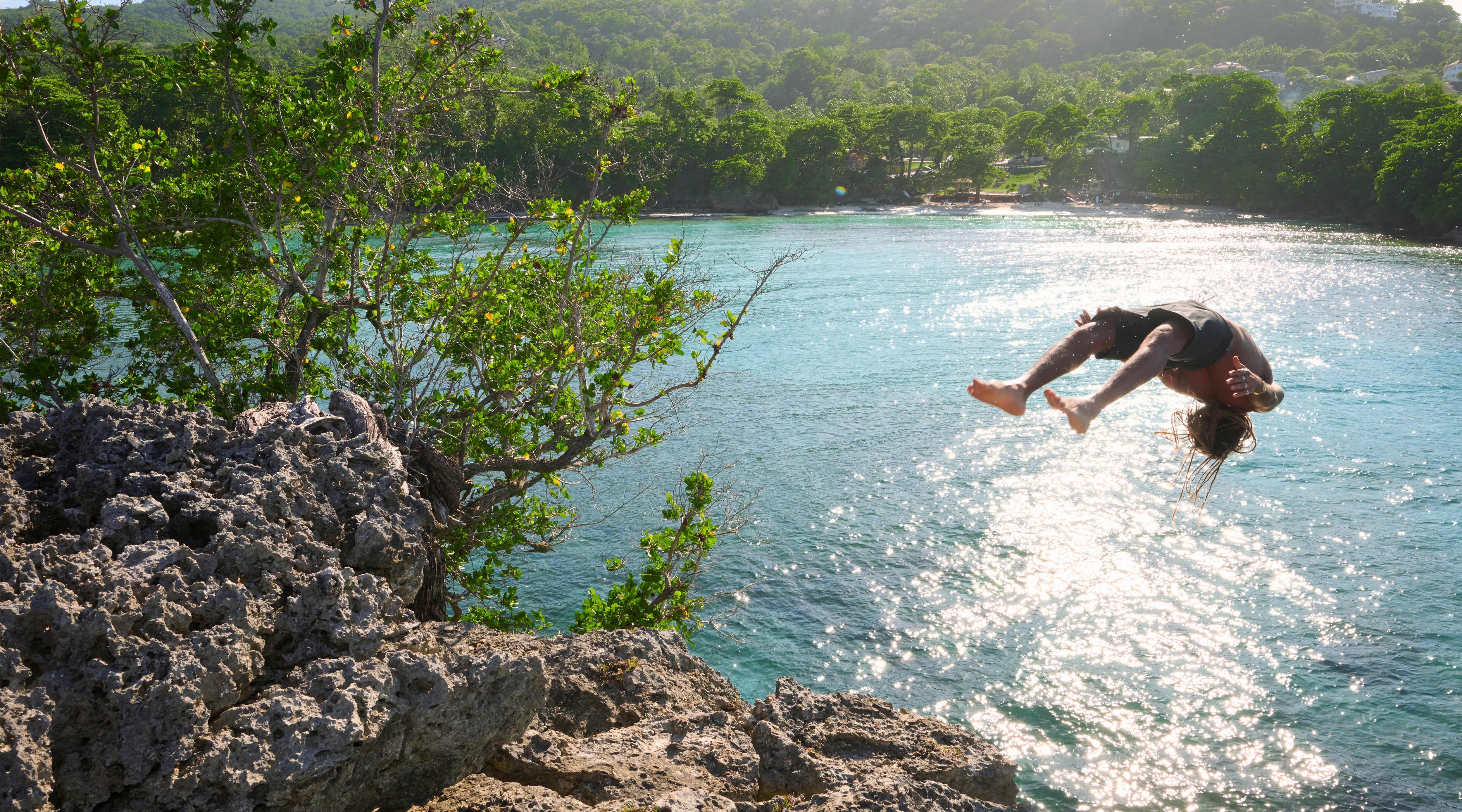 Person jumping off a cliff into clear blue water with lush greenery in the background