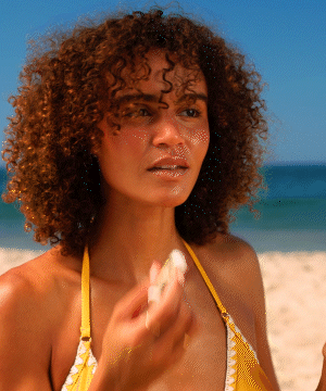 A person with natural curly hair applying Original SPF 30 Sunscreen Lip Balm on a sunny beach, wearing a yellow swimsuit top, demonstrating sun protection for lips - Sun Bum Original SPF 30 Sunscreen Lip Balm 3-Pack.