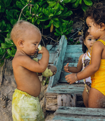 Toddler applying sunscreen stick while holding a plush toy, surrounded by friends at the beach, from the SB Baby Sunscreen Collection – Sun Bum.