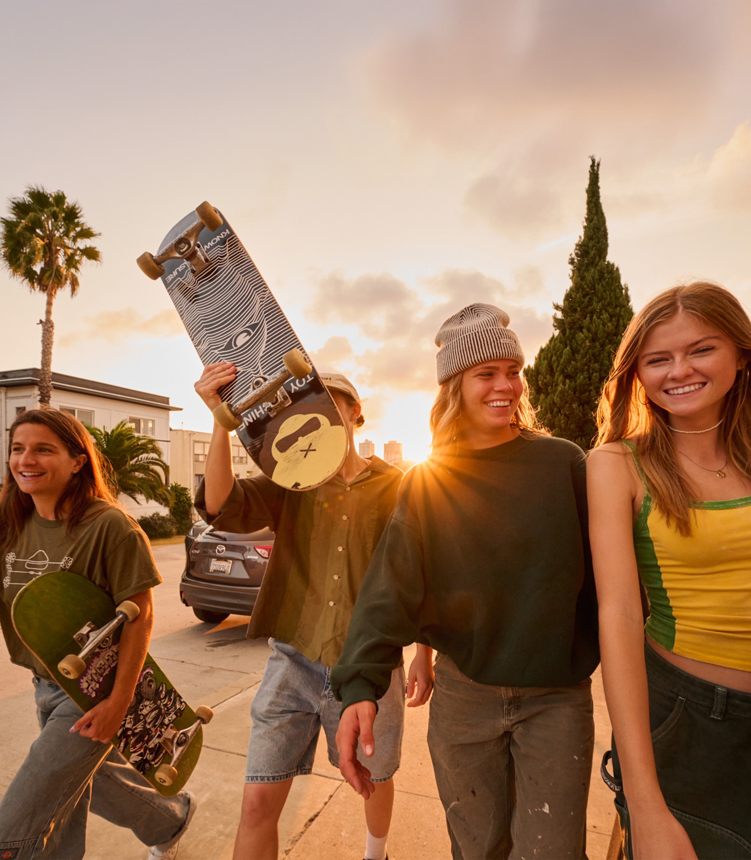 Four friends smiling and skating at sunset, holding boards from the SB Sun Care Collection – Sun Bum.