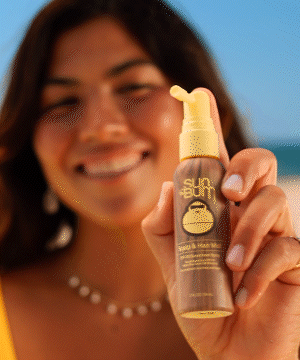 A person holding up Sun Bum Scalp & Hair Mist SPF 30 at the beach, displaying the golden-colored spray bottle against a blue sky background. The lightweight sun protection spray promises non-greasy coverage for hair and scalp - by Sun Bum