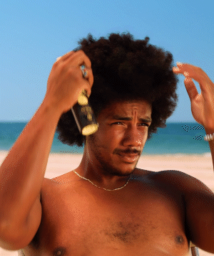 A person at the beach spraying Sea Spray hair texturizer, demonstrating the beach-inspired styling product against a scenic ocean backdrop. Natural curly hair texture shown being enhanced with Sun Bum Sea Spray.