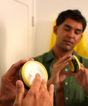 A person holding and examining a jar of Texturizing Surf Paste against a yellow background while demonstrating the product's texture and application. Perfect for achieving that effortless beach-styled hair look - by Sun Bum