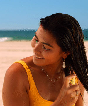 A person with wet, dark hair wearing a yellow tank top applies Sun Bum x Tangle Teezer Ultimate Detangling Duo hair product at the beach, smiling with ocean waves and sand in the background - by Sun Bum
