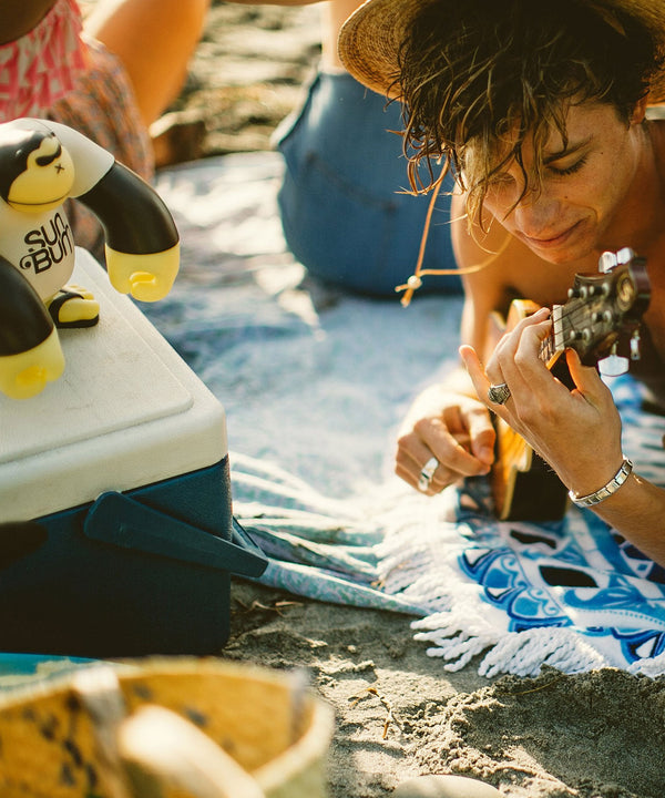 "Person playing guitar at the beach with a 'Sonny' 9" Vinyl Figure on a cooler, Sun Bum"