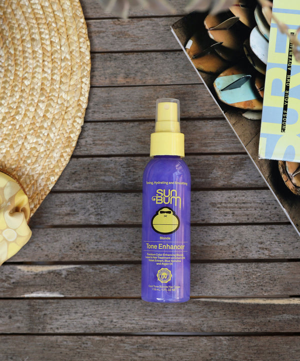Blonde Tone Enhancer by Sun Bum, featuring a purple bottle with yellow cap on a wooden surface with a straw hat and a magazine nearby, aimed to reduce yellow and orange tones using Violet Extract & Argan Oil.