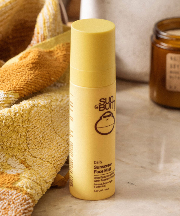 Yellow bottle of Sun Bum Daily Sunscreen Face Mist SPF 30 on a bathroom counter, surrounded by towels and a candle.