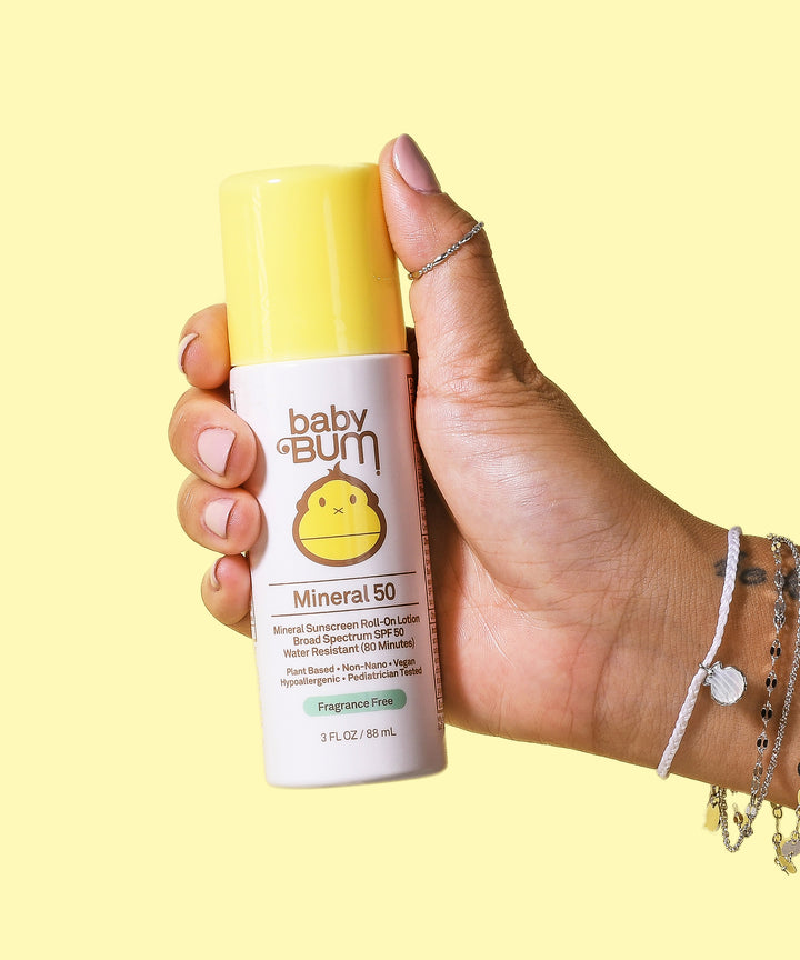 Hand holding Baby Bum Mineral SPF 50 Roll-On Sunscreen bottle against a light yellow background. Baby Bum,