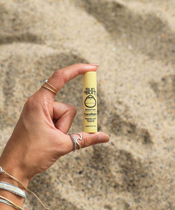 Hand holding Sun Bum CocoBalm Lip Balm in Banana Cream flavor against a sandy beach background, Sun Bum.