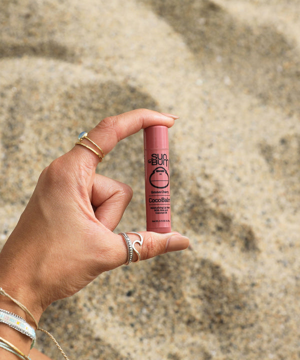 Hand holding CocoBalm Lip Balm - Groove Cherry against sandy background, Sun Bum.