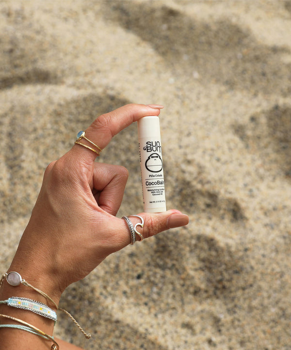 Hand holding CocoBalm Lip Balm - Piña Colada by Sun Bum in front of sandy beach background