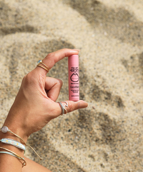 Hand holding a pink tube of CocoBalm Lip Balm - Wild Strawberry flavor, Sun Bum, against a sandy beach background.