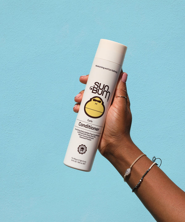 Hand holding a bottle of Curls Conditioner against a blue background, featuring Kukui Nut Oil and Monoi Coconut Oil for enhanced curls. Sun Bum.