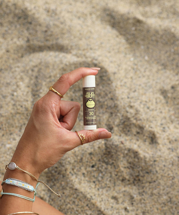 Hand holding Original SPF 30 Sunscreen Lip Balm - Coconut at the beach, offering Aloe and Vitamin E protection. Sun Bum