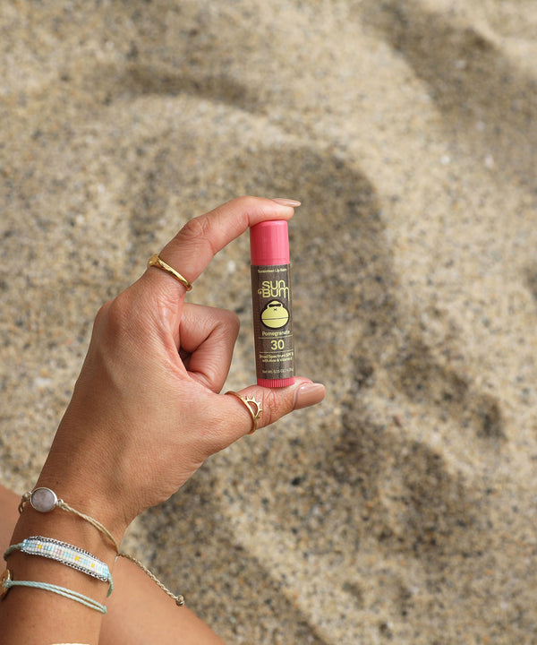 Hand holding Original SPF 30 Sunscreen Lip Balm - Pomegranate at the beach; Aloe and Vitamin E-infused for all-day protection and moisturization, Sun Bum.