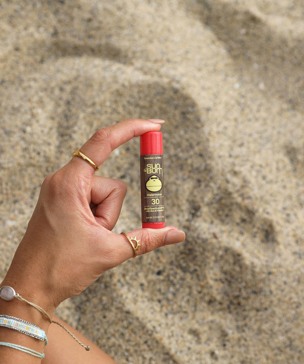 Hand holding Original SPF 30 Sunscreen Lip Balm in Watermelon flavor against a sandy beach background. Sun Bum.