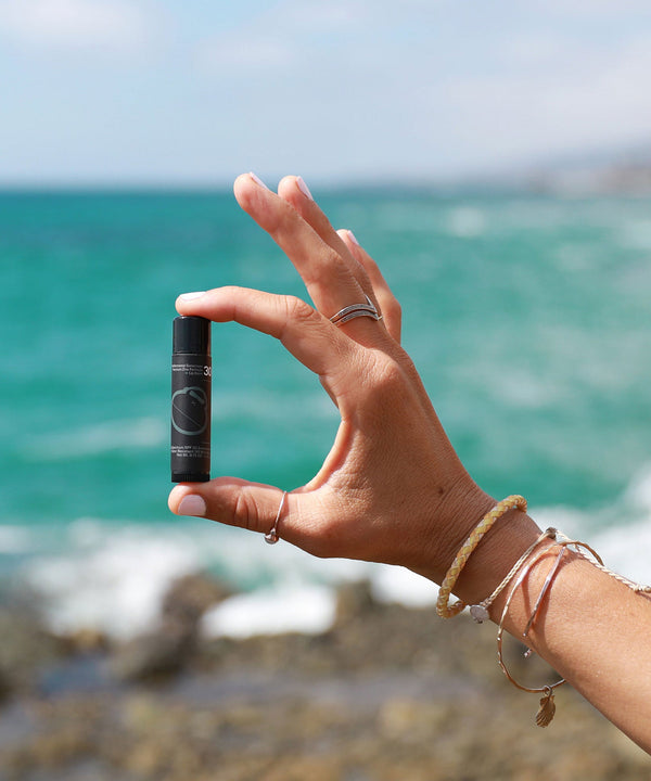 Hand holding Signature SPF 30 Sunscreen Lip Balm with ocean background, perfect for sun protection outdoors. Sun Bum.