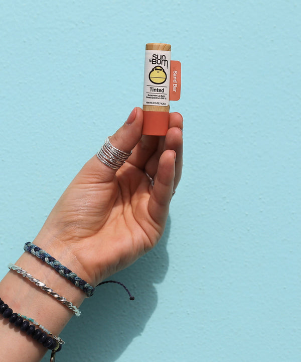 Hand holding a tube of Tinted SPF 15 Lip Balm - Sand Bar against a light blue background. Sun Bum