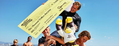 Four men on a beach holding a large yellow check and a cartoon mascot figure. One man wears a black wetsuit, and the background shows blue sky and ocean.