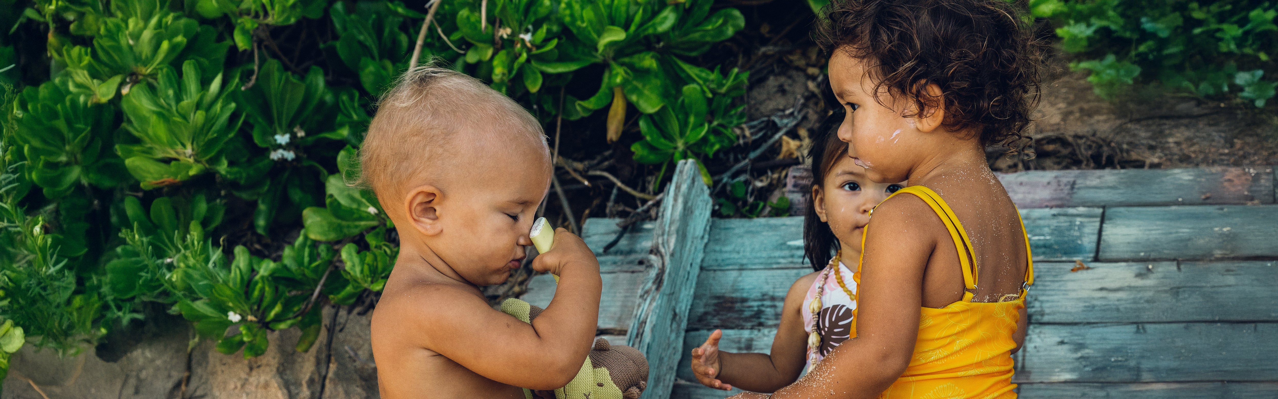 Toddler applying sunscreen stick while holding a plush toy, surrounded by friends at the beach, from the SB Baby Sunscreen Collection – Sun Bum.
