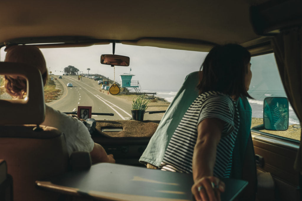 View from inside a van as two people travel along a coastal road with a beach and lifeguard tower outside
