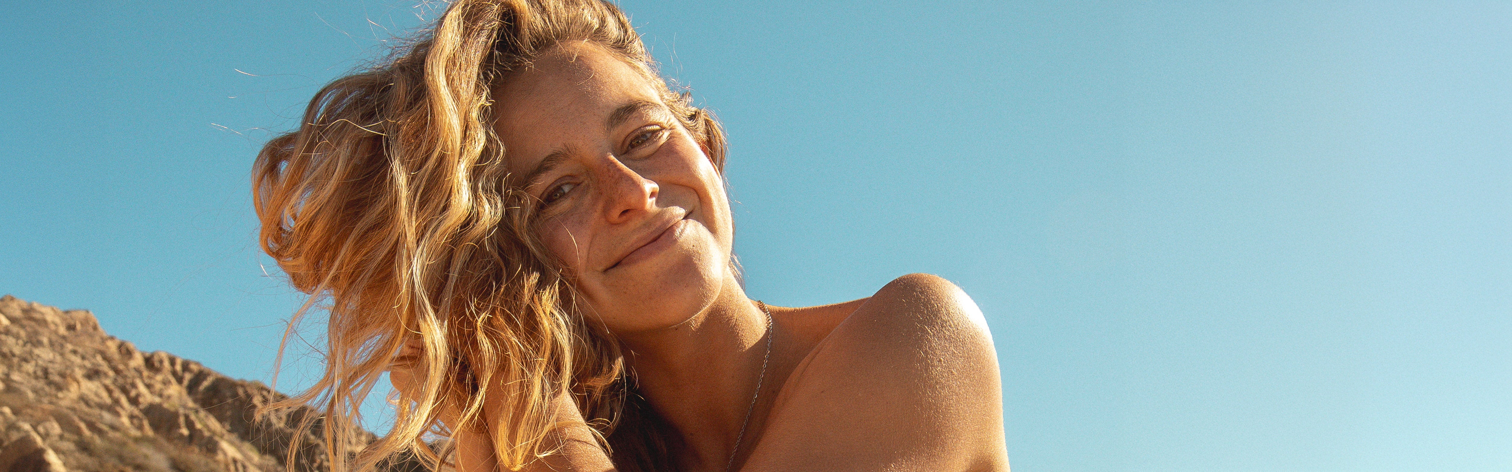 Smiling woman with sun-kissed blonde waves outdoors, highlighting the SB Blonde Hair Care Collection – Sun Bum.