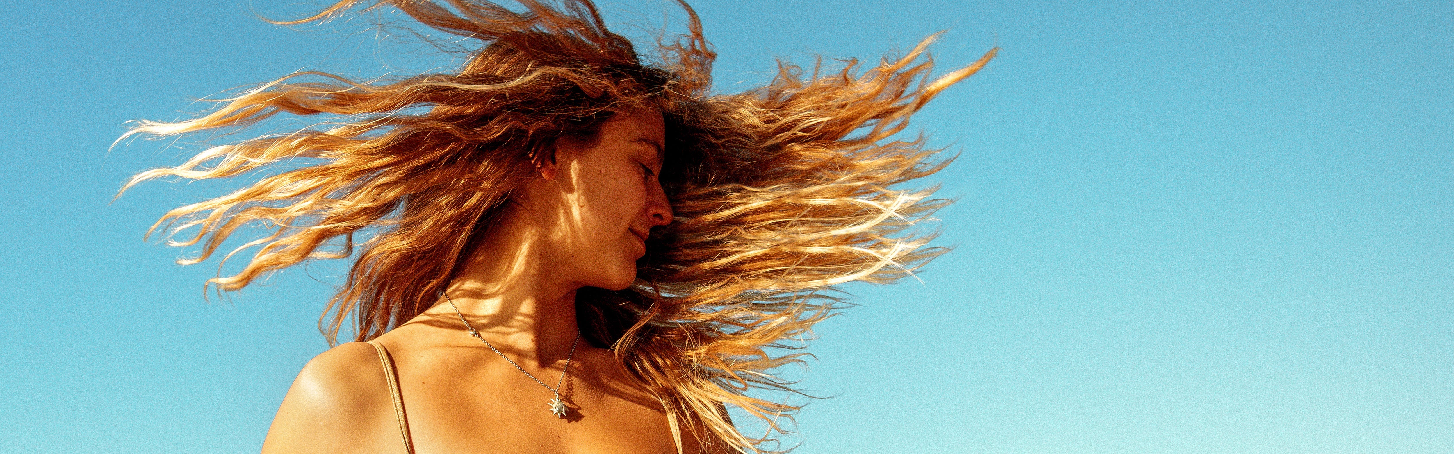 Woman flipping her sunlit, beachy hair under a clear sky, featuring the SB Revitalizing Hair Care Collection – Sun Bum.