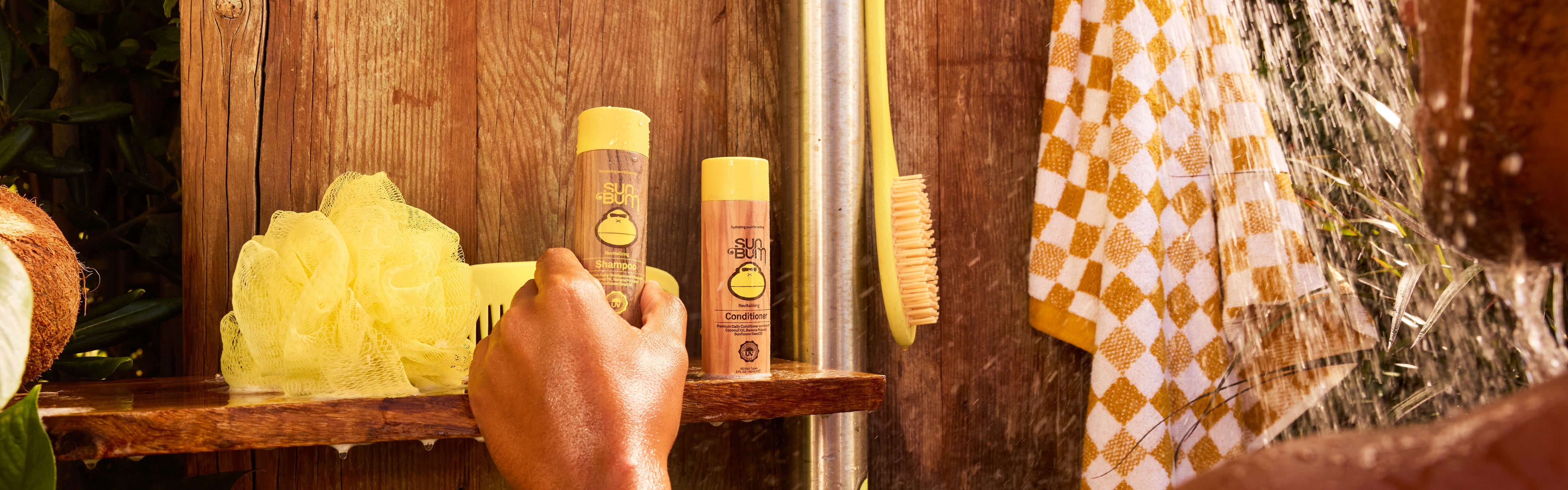 Wet hand reaching for mini Sun Bum shampoo and conditioner bottles on a wooden shower shelf – Travel Hair Care Collection - Sun Bum.