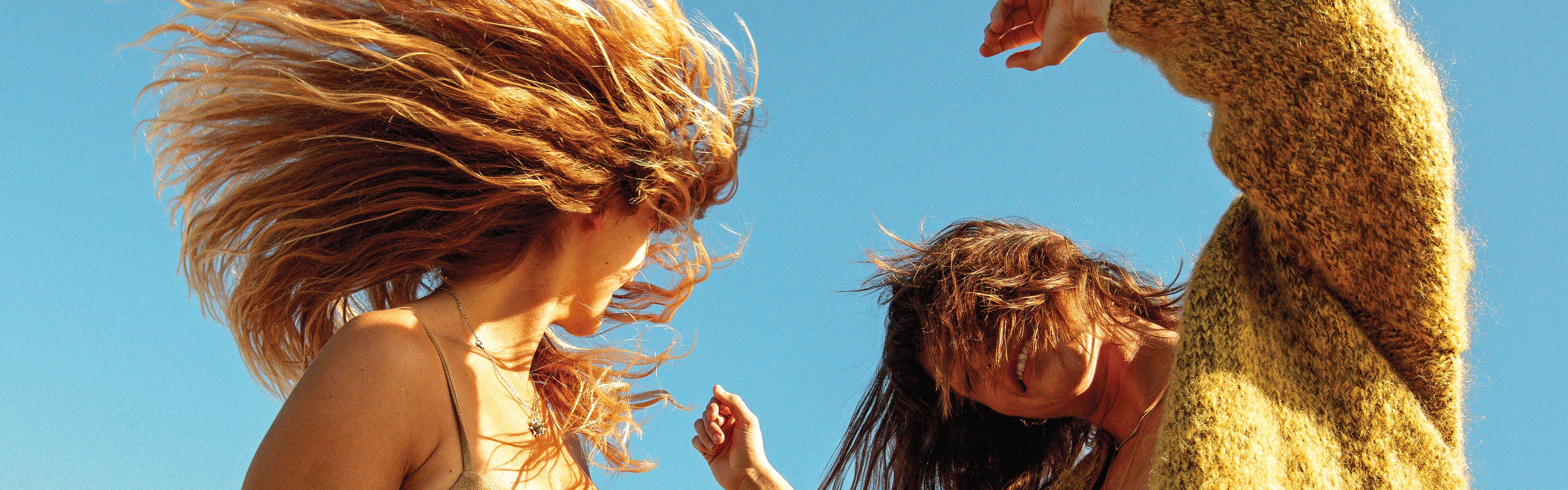 Two women dancing outdoors with textured, wind-blown hair under the sun, featuring the SB Texturizing Hair Care Collection – Sun Bum.