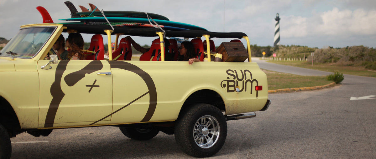 Yellow Sun Bum truck carrying multiple surfboards parked on a road near a lighthouse with cloudy sky above.